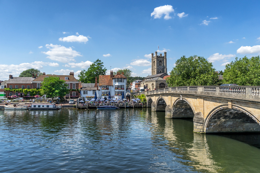 View of Henley-on-Thames, Henley bridge over the Thames on a bright day. Property along the river, a pub with many patrons outside, boats in the river. 