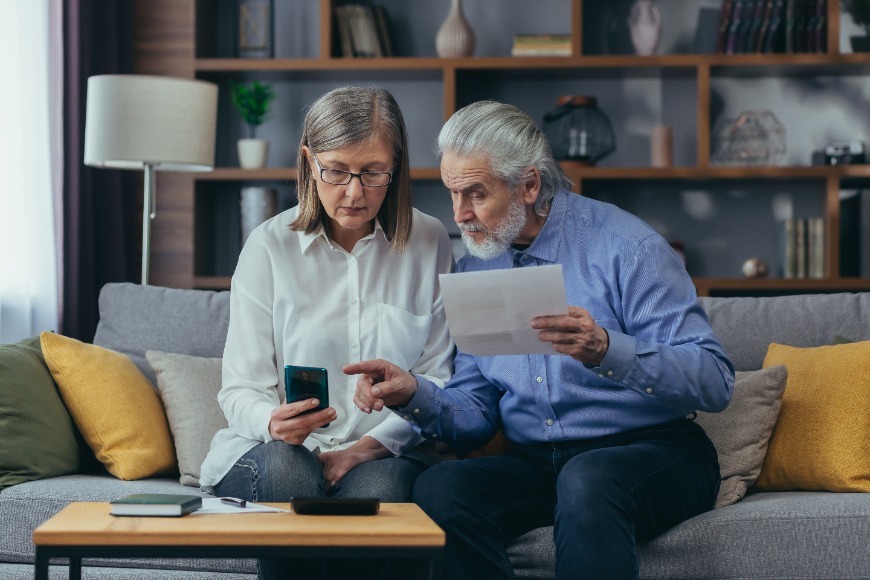 Male and female pensioners sat next to each other on a sofa reviewing a document and something on a smart phone. A small table is in front of them with a book and a pen and paper.
